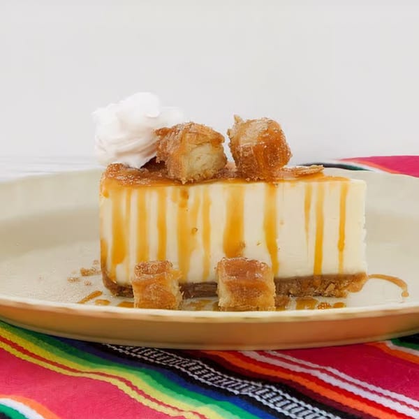 Cheesecake slice with caramel drizzle, whipped cream, and churro pieces on a colorful Mexican blanket.