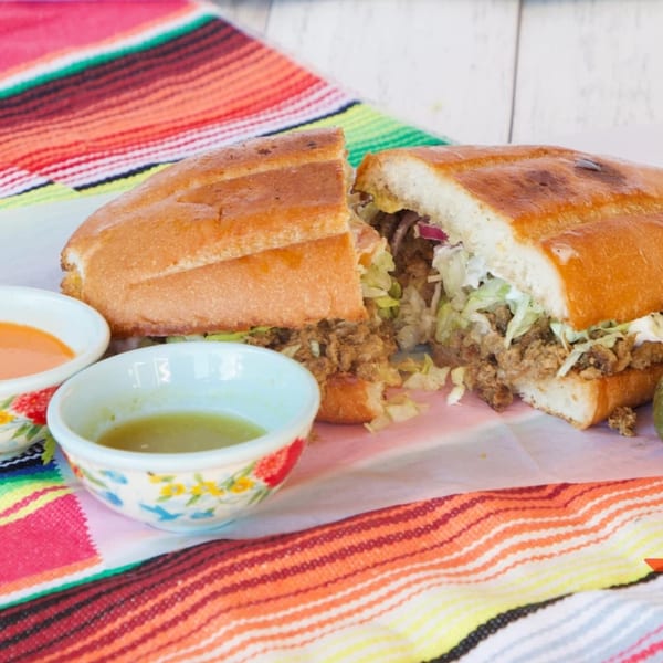 A large Mexican torta sandwich, cut in half, with two small bowls of sauce on a colorful striped cloth.