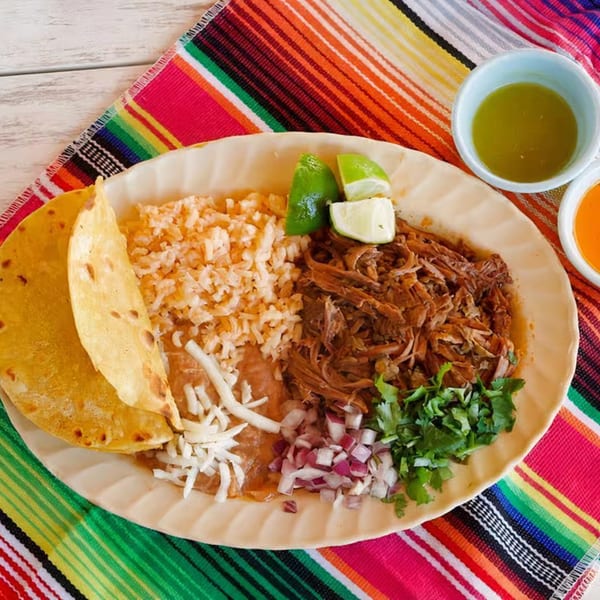 A Mexican dish with shredded meat, rice, beans, tortillas, lime, cilantro, and onions on a colorful placemat.